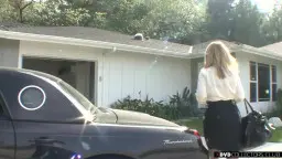 Blonde Stiefmutter in weißer Bluse und schwarzem Rock steht vor ihrem Haus neben einem schwarzen Auto.