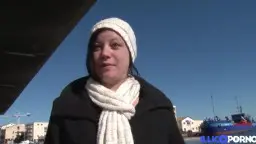 Mollige Milf mit weißer Mütze und dickem Schal steht lächelnd am sonnigen Hafen.